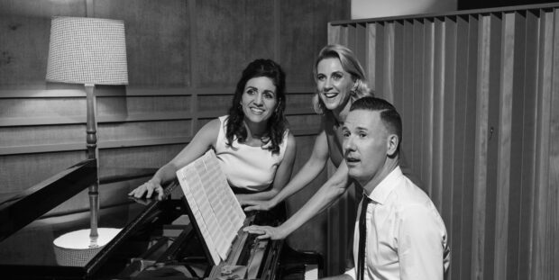 Black and white, 1950's stylised photo of three people smiling around a piano.