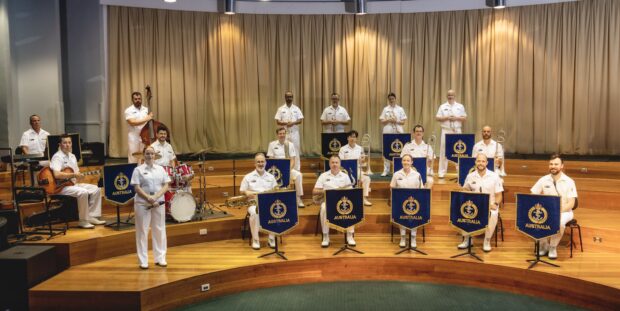 The Royal Australian Navy Band members in white uniforms, stages on a gold backdropped stage.