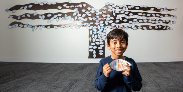 A smiling child standing infront of a the Message Tree installation with a paper leave.