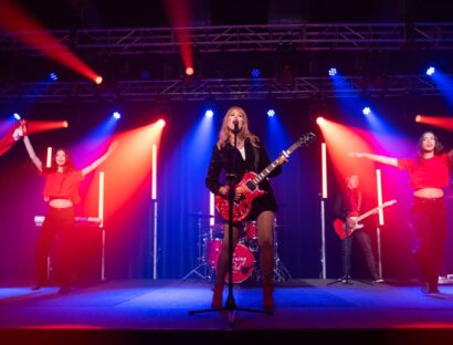 Blonder female performer on stage with a guitar and two backup dancers and a band, with blue and red lighting.